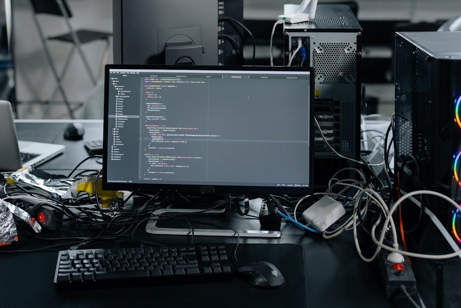 A black laptop with a red backlit keyboard is open on a dark surface, displaying a blurred screen with lines of code or text, indicating a technical or digital environment. Next to the laptop, there i
