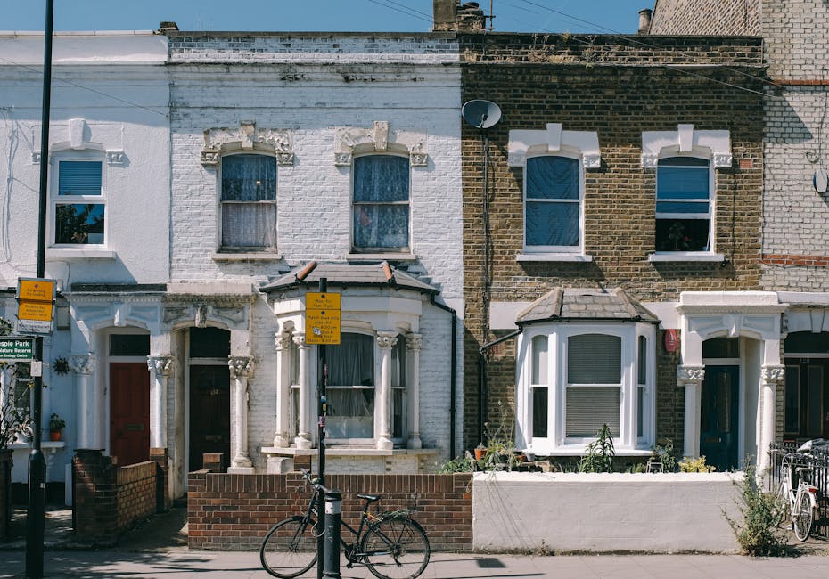 Photograph of a residential street in Brixton SW2 featuring a row of traditional terraced houses with brick facades and bay windows, some with open windows and window boxes. The street is lined with p