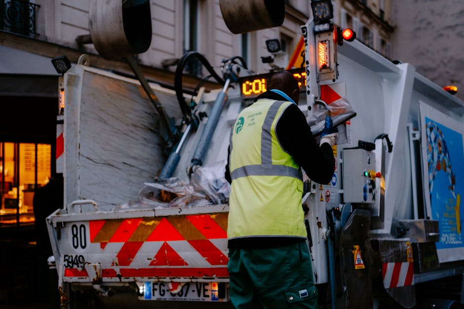 A waste collection truck parked on a narrow cobblestone street in an urban area, with a worker wearing a blue uniform, high-visibility orange vest, and blue cap, standing beside the truck and emptying