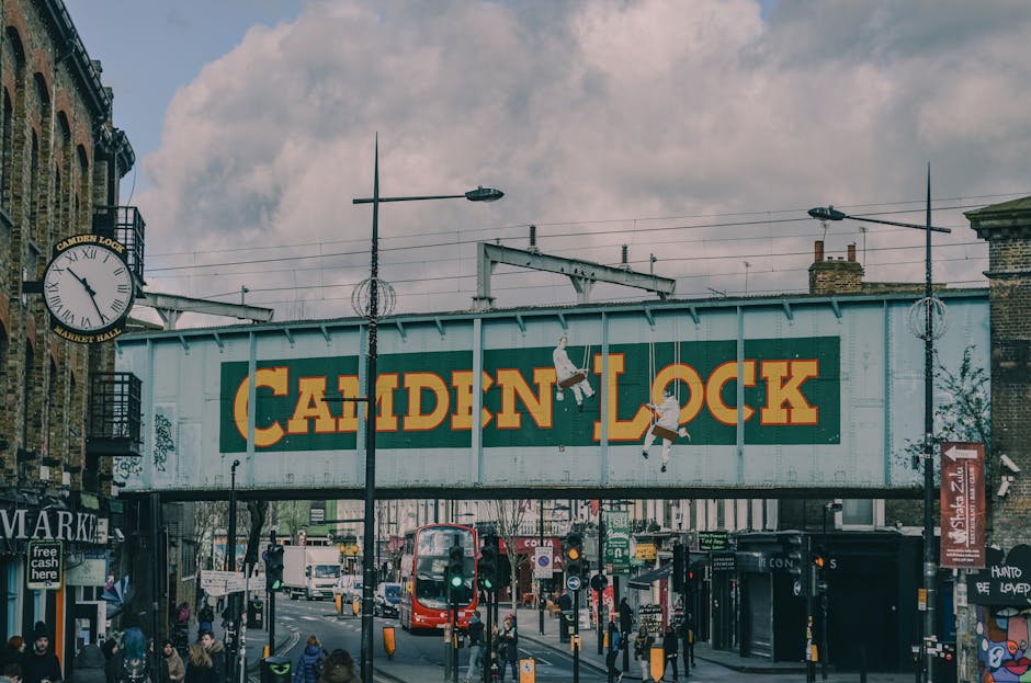 A large, weathered metal railway bridge painted in light blue spans across a busy street in Camden, London. The bridge features a prominent yellow sign with the words 'CAMDEN LOCK' in bold letters, ac