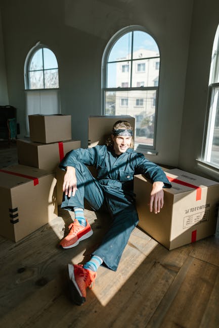 A young woman sitting on the floor inside a room surrounded by large cardboard boxes, some sealed with red packing tape and others open, revealing packing materials like white foam packing peanuts. Sh